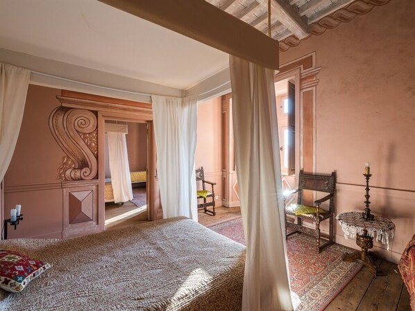 Un dormitorio con una cama grande, una silla, una mesa con una vela y vistas a otra habitación por una puerta abierta.