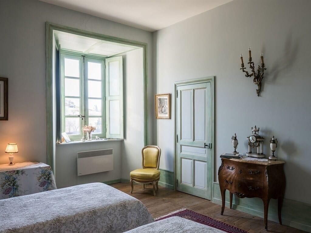 A room with two beds, a chair, a dresser, a window with shutters, and a wall-mounted candelabra.