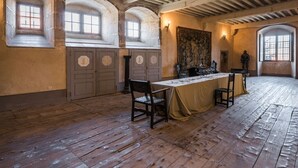 Una habitación con vigas de madera, ventanas arqueadas, una mesa con sillas y un tapiz.