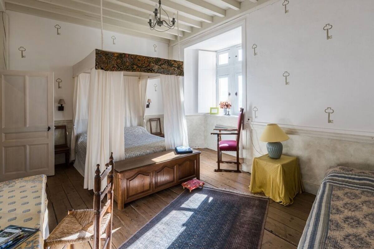 A bedroom with a canopy bed, a wooden dresser, a chair, a small table with a lamp, and a window with curtains.