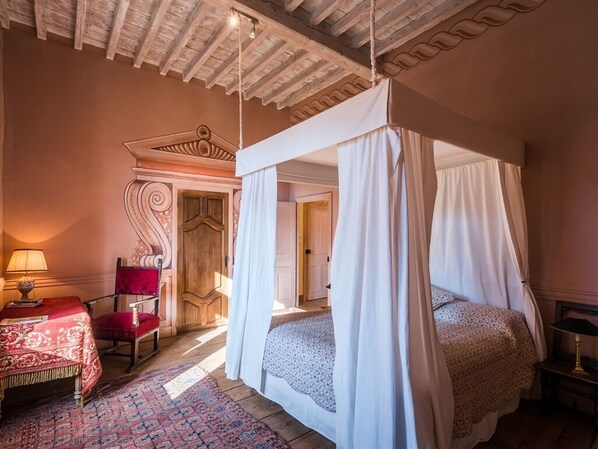 Ein Schlafzimmer mit einem Himmelsbett, einem roten Sessel, einem Tisch mit Lampe und einem gemusterten Teppich.