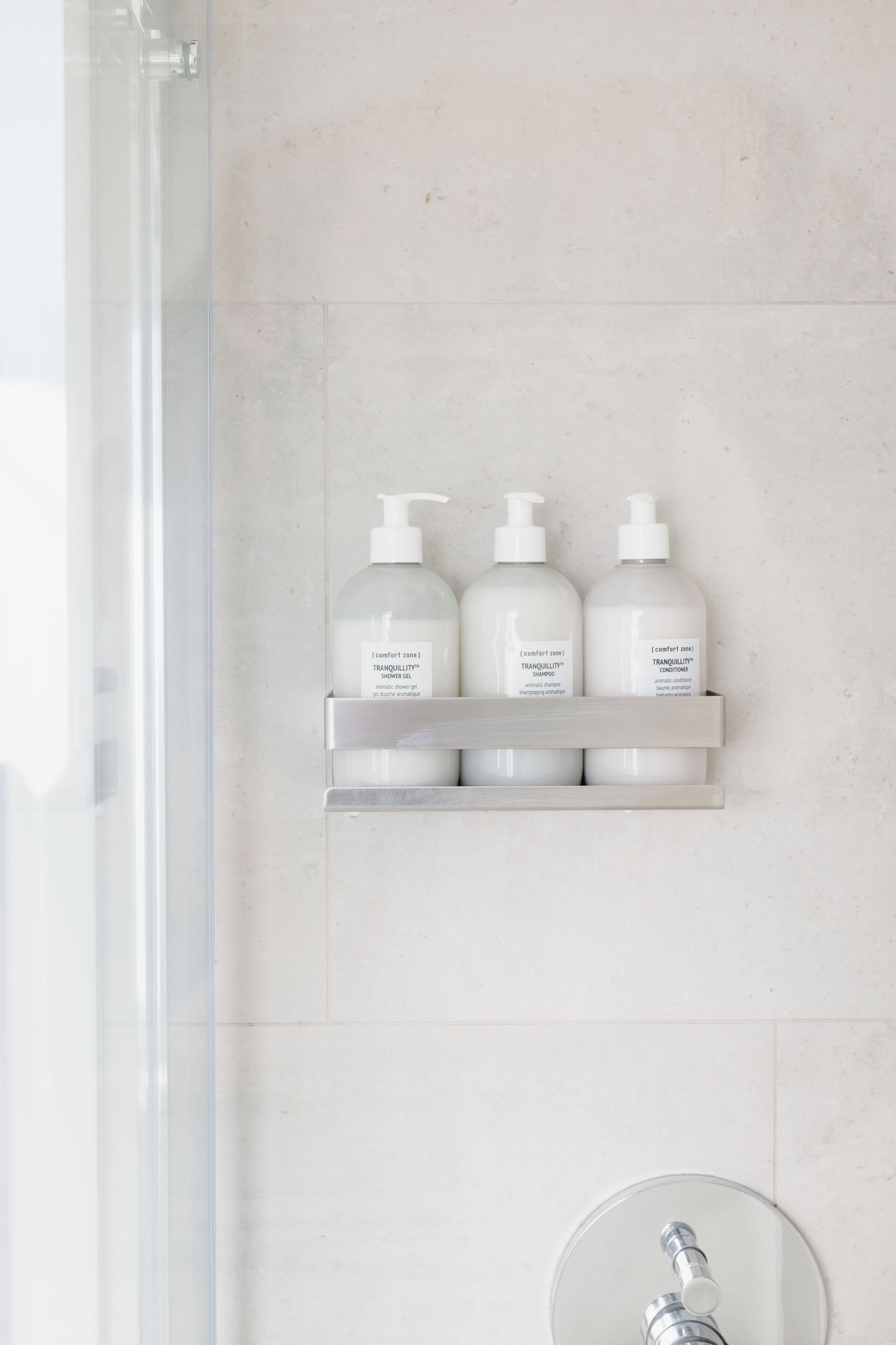 Un baño con mampara de vidrio, un lavabo blanco y un estante con tres botellas de gel de ducha, champú y acondicionador 'Tranquility'.