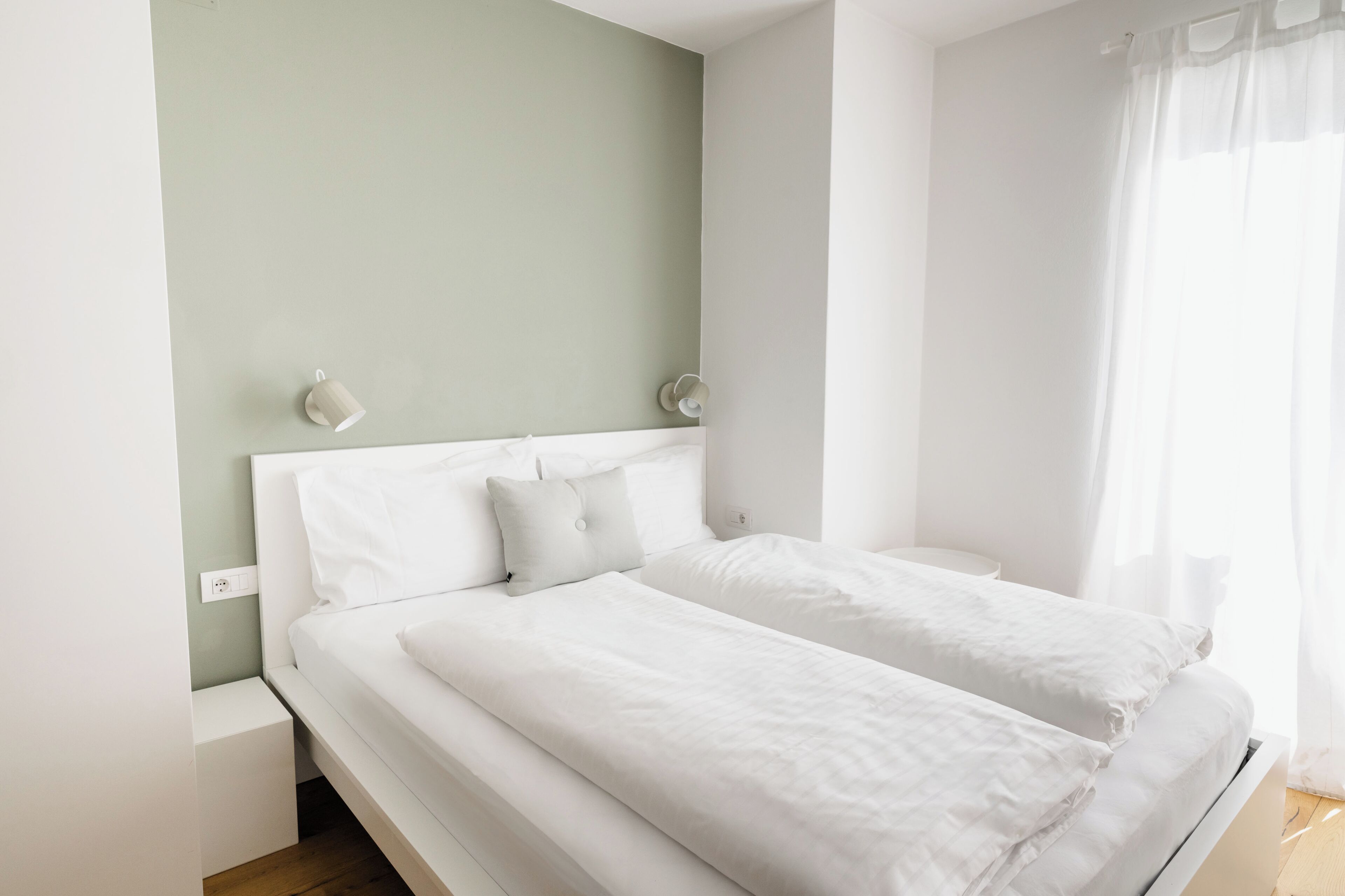 Una cama doble con ropa blanca, una pared verde claro y una mesita de noche blanca.