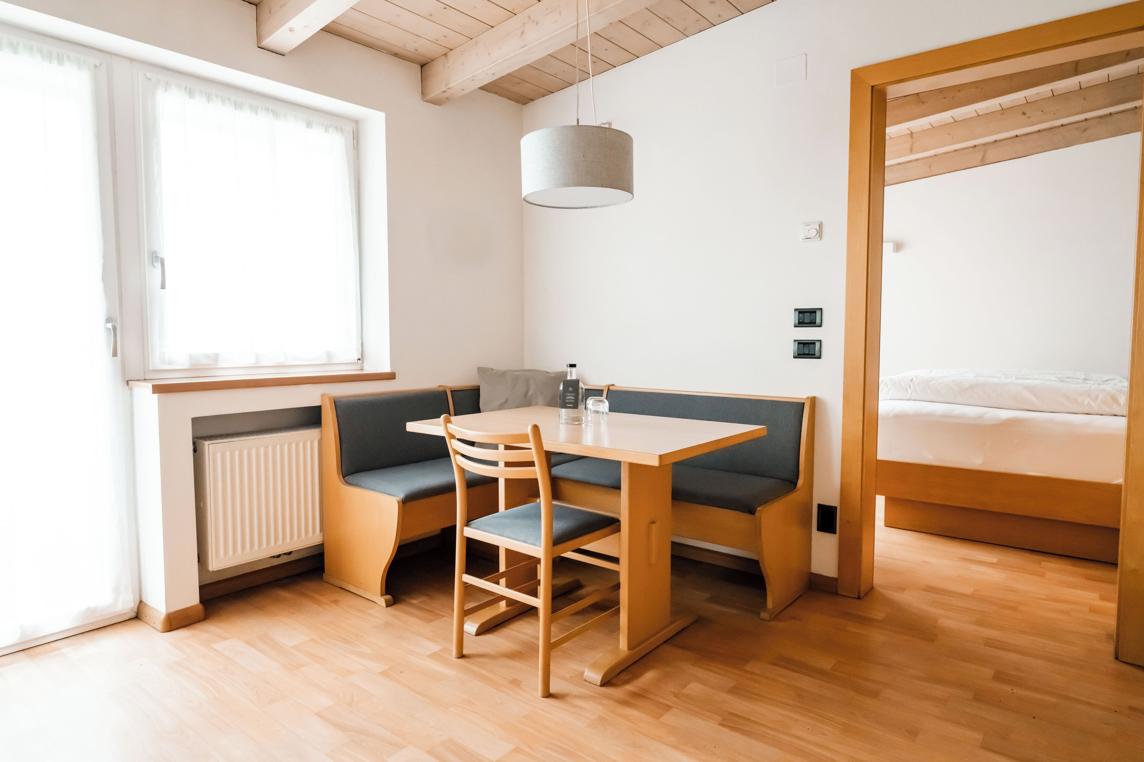 Una habitación con una mesa de madera y un banco, una cama, un radiador y una ventana.