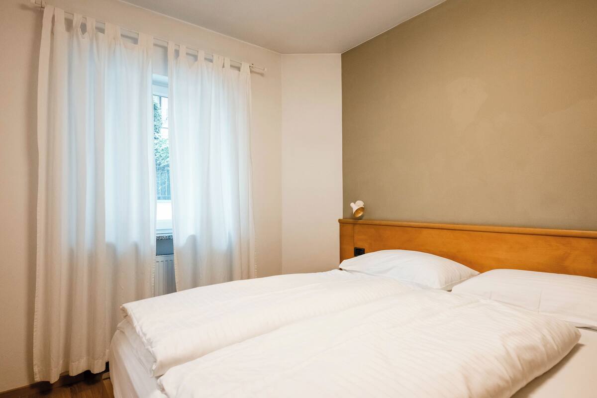 A hotel room with a bed, white curtains, a wooden headboard, and a view through the window.