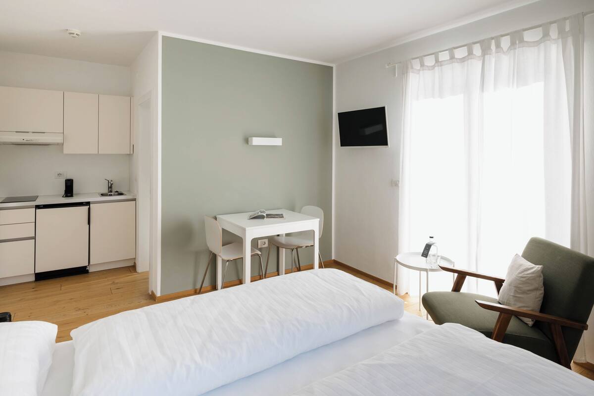 A hotel room with a bed, a desk, a chair, a small table, a TV, and a kitchenette.