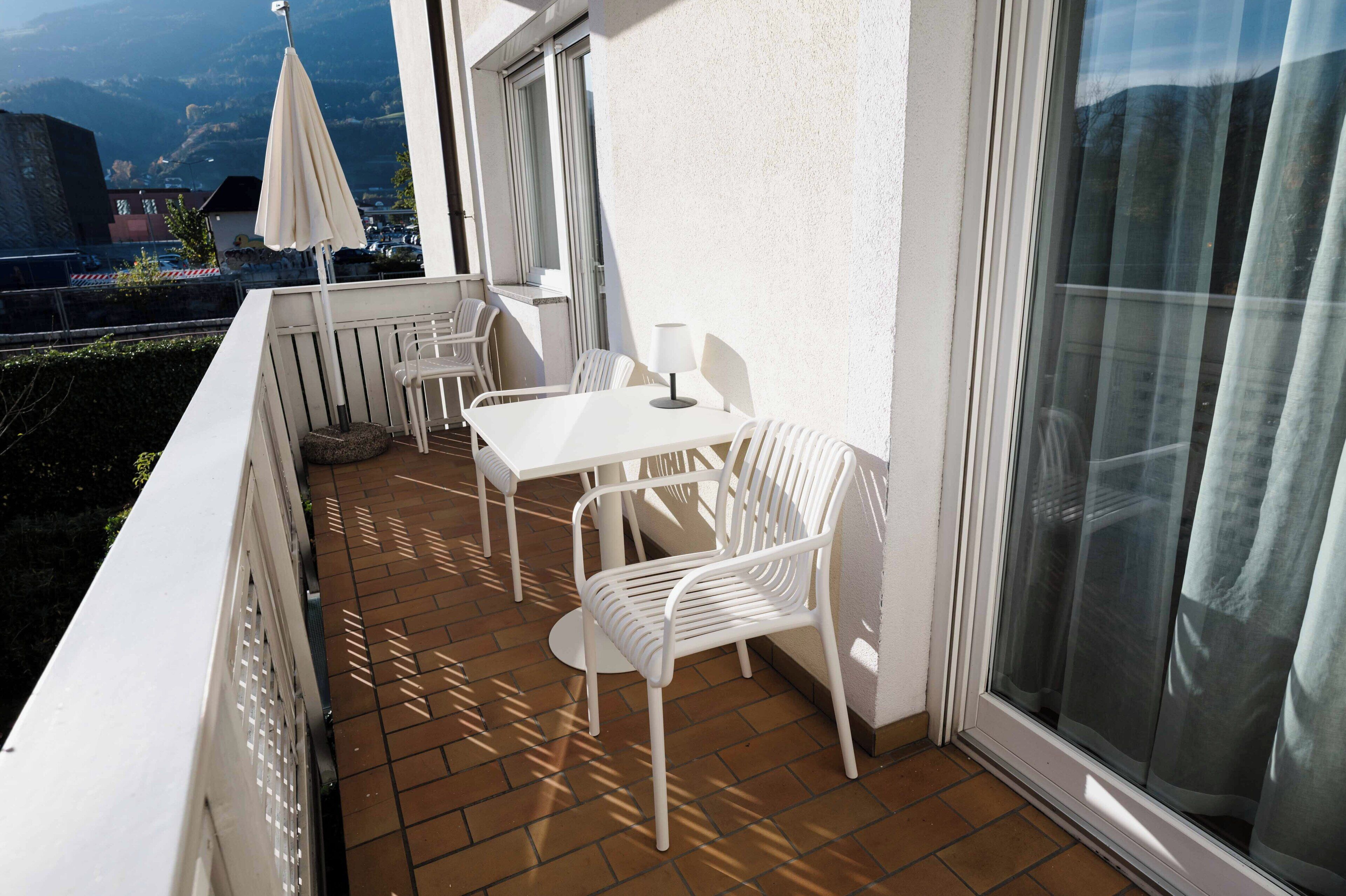 A balcony with a white table, two white chairs, a white umbrella, and a window with a curtain.