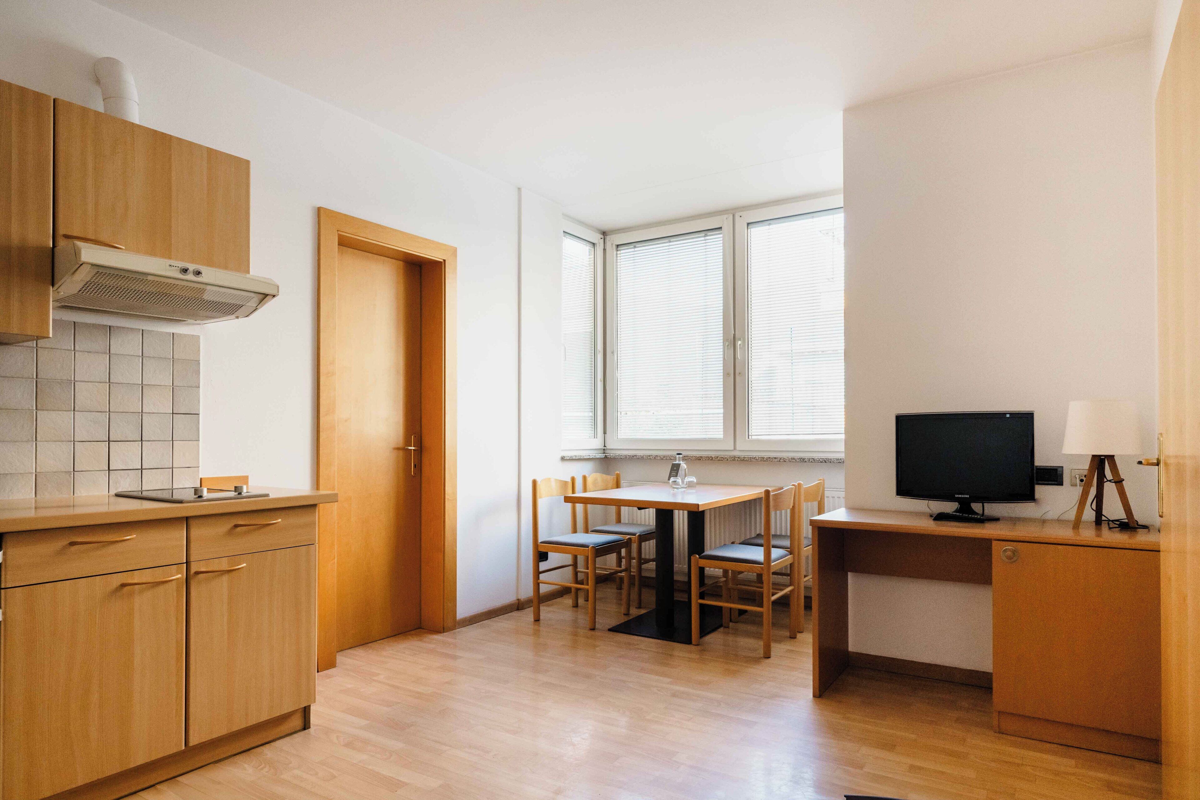 Una cocina con armarios de madera, salpicadero de azulejos, una mesa de comedor con sillas, un televisor sobre un escritorio y una ventana con persianas.