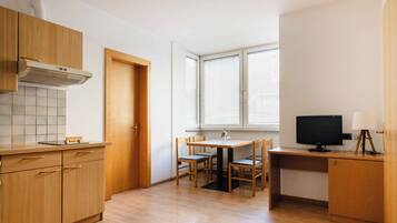 Una cocina con armarios de madera, salpicadero de azulejos, una mesa de comedor con sillas, un televisor sobre un escritorio y una ventana con persianas.