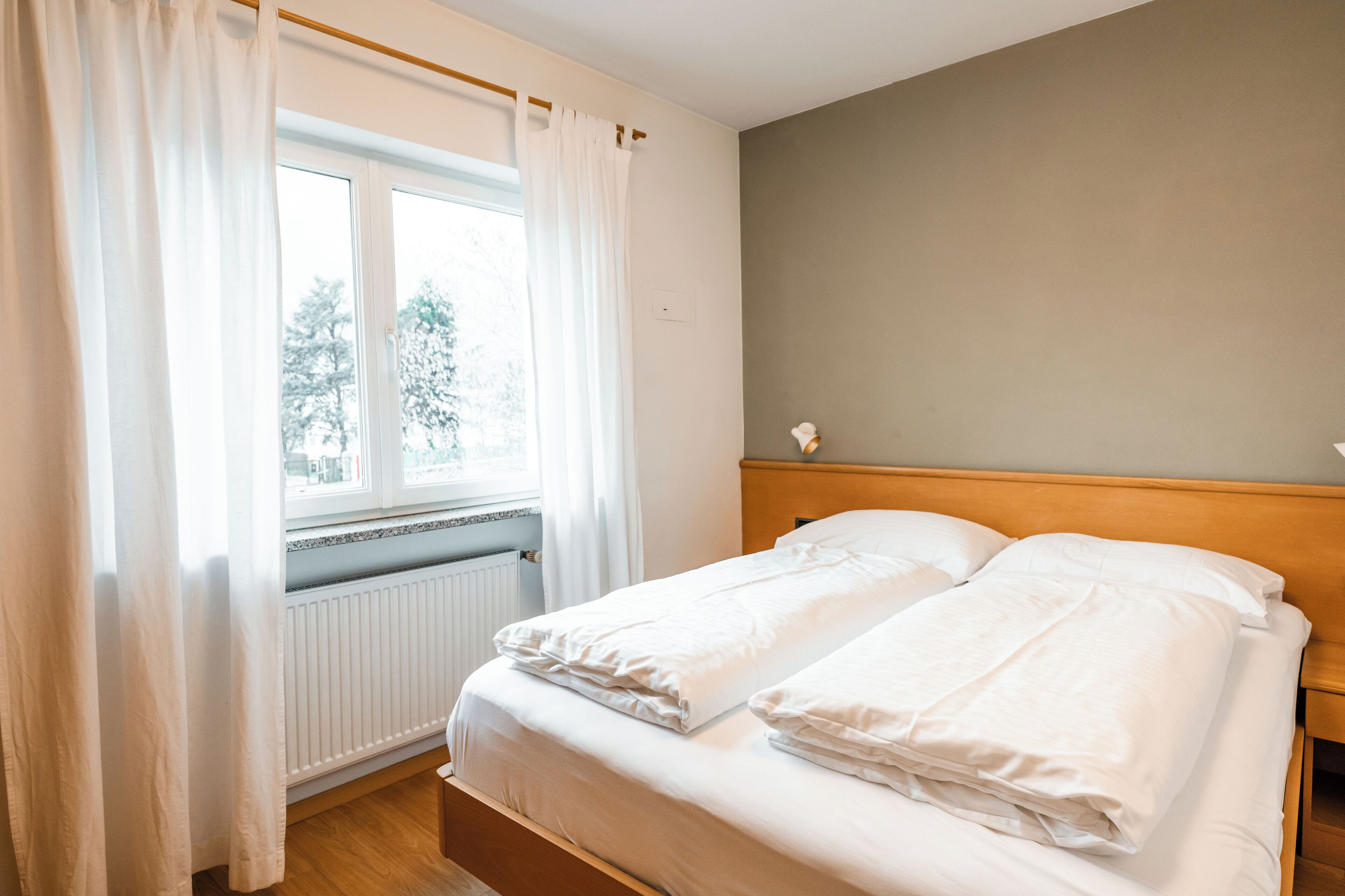 Un dormitorio con cama doble, ropa de cama blanca, cabecera de madera, ventana con cortinas blancas y vista a árboles.