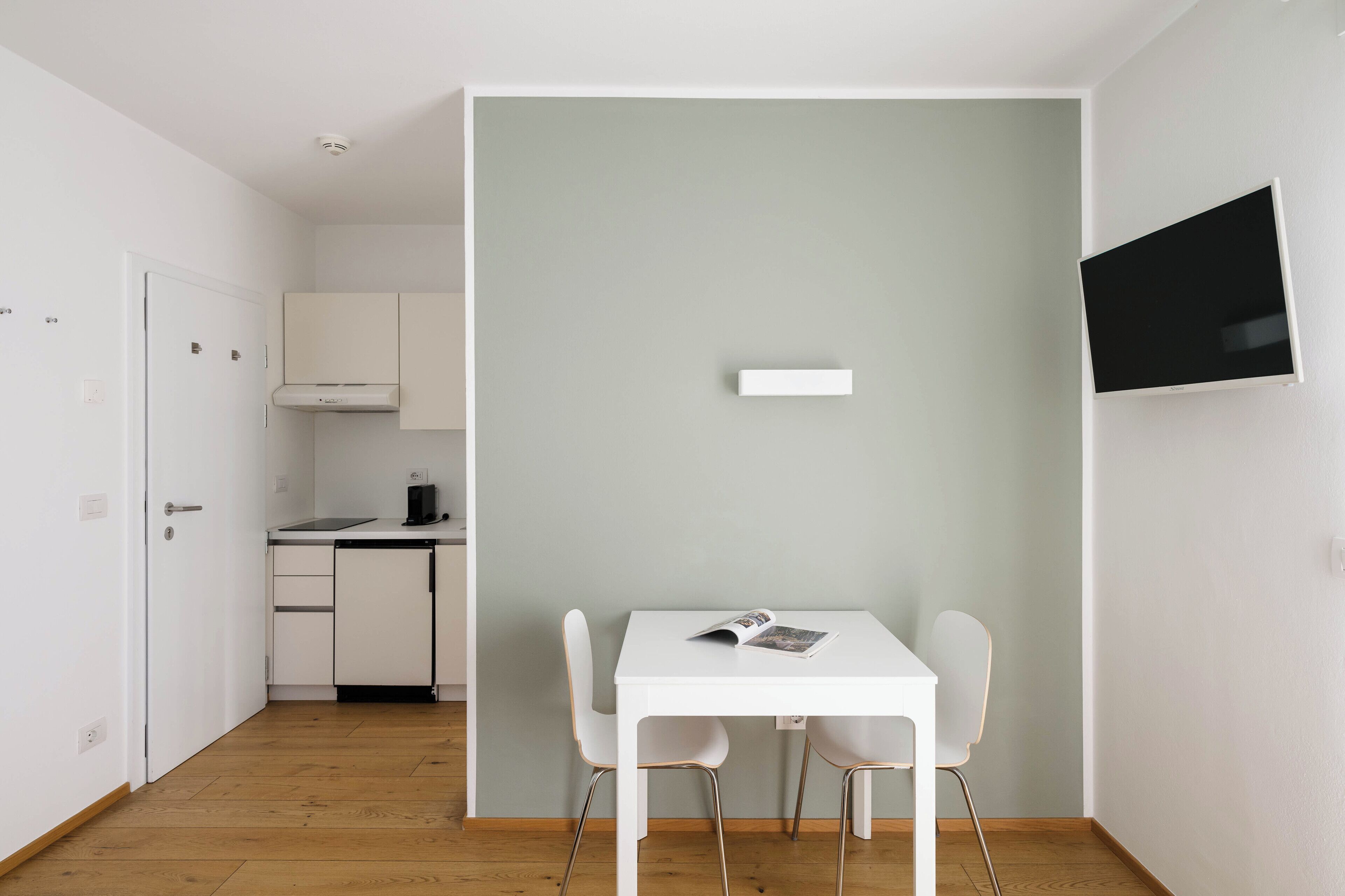Eine kleine Küche mit weißen Schränken, einem weißen Tisch mit zwei Stühlen und einem an der Wand befestigten Fernseher.