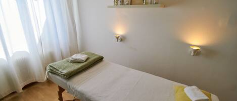 A single bed with a green towel and a yellow pillow, a wooden floor, a white curtain, and a shelf with toiletries.