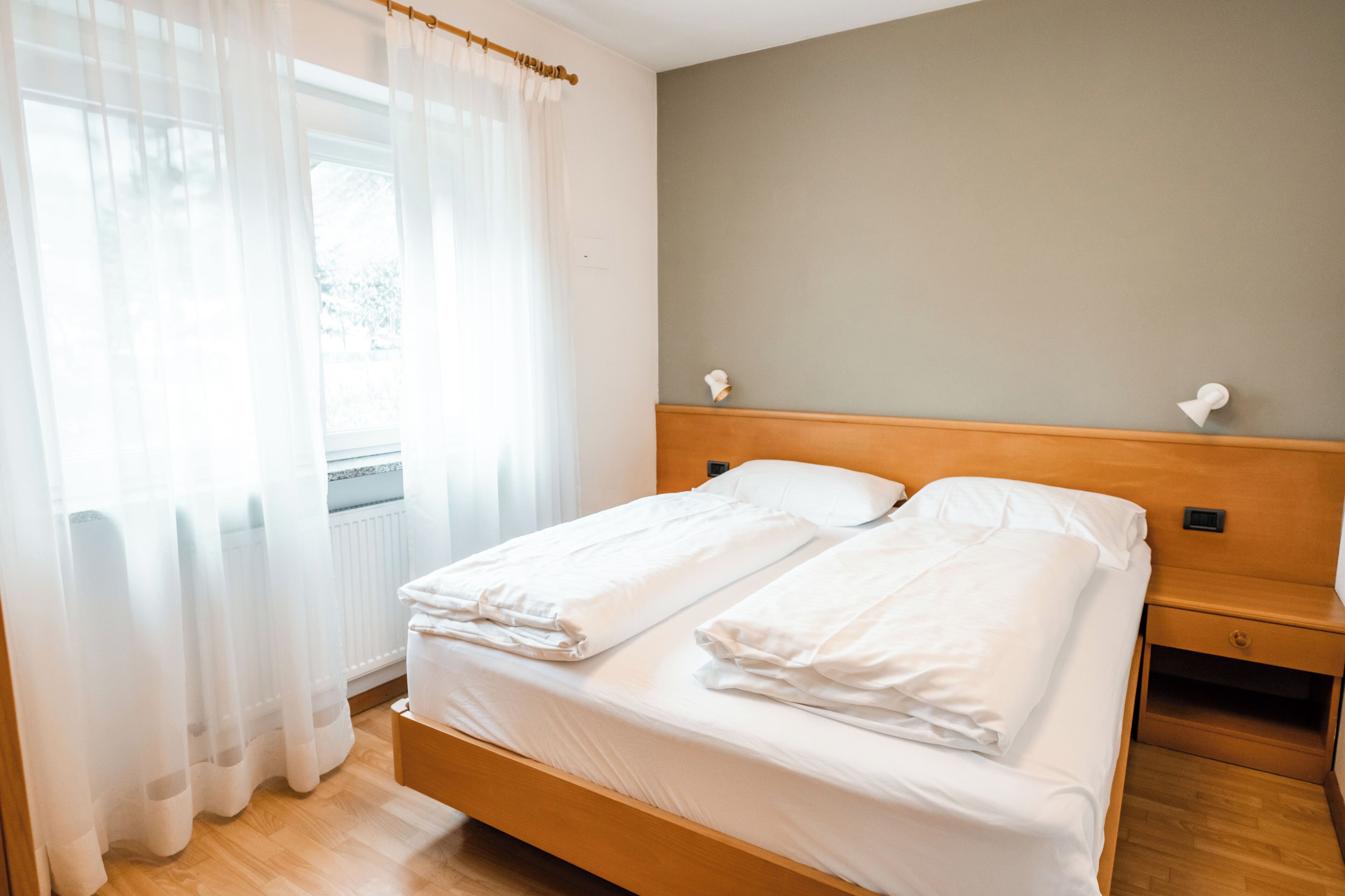 A double bed with white linens, a wooden headboard, a bedside table with a lamp, and sheer curtains on a window.