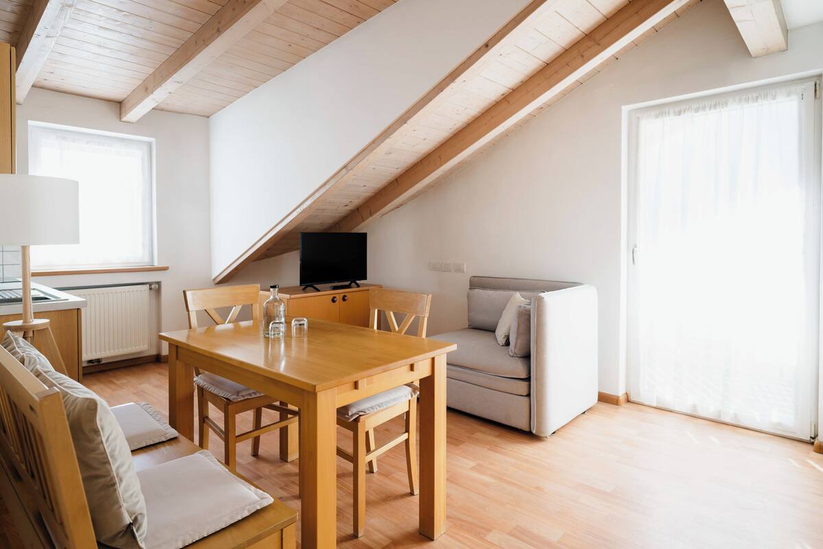 A modern loft apartment with a dining area, a sofa, a television, and a large window.