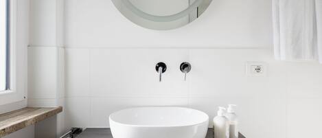 A modern bathroom with a large white sink, a round mirror, and two faucets. There are two bottles of toiletries on the counter.