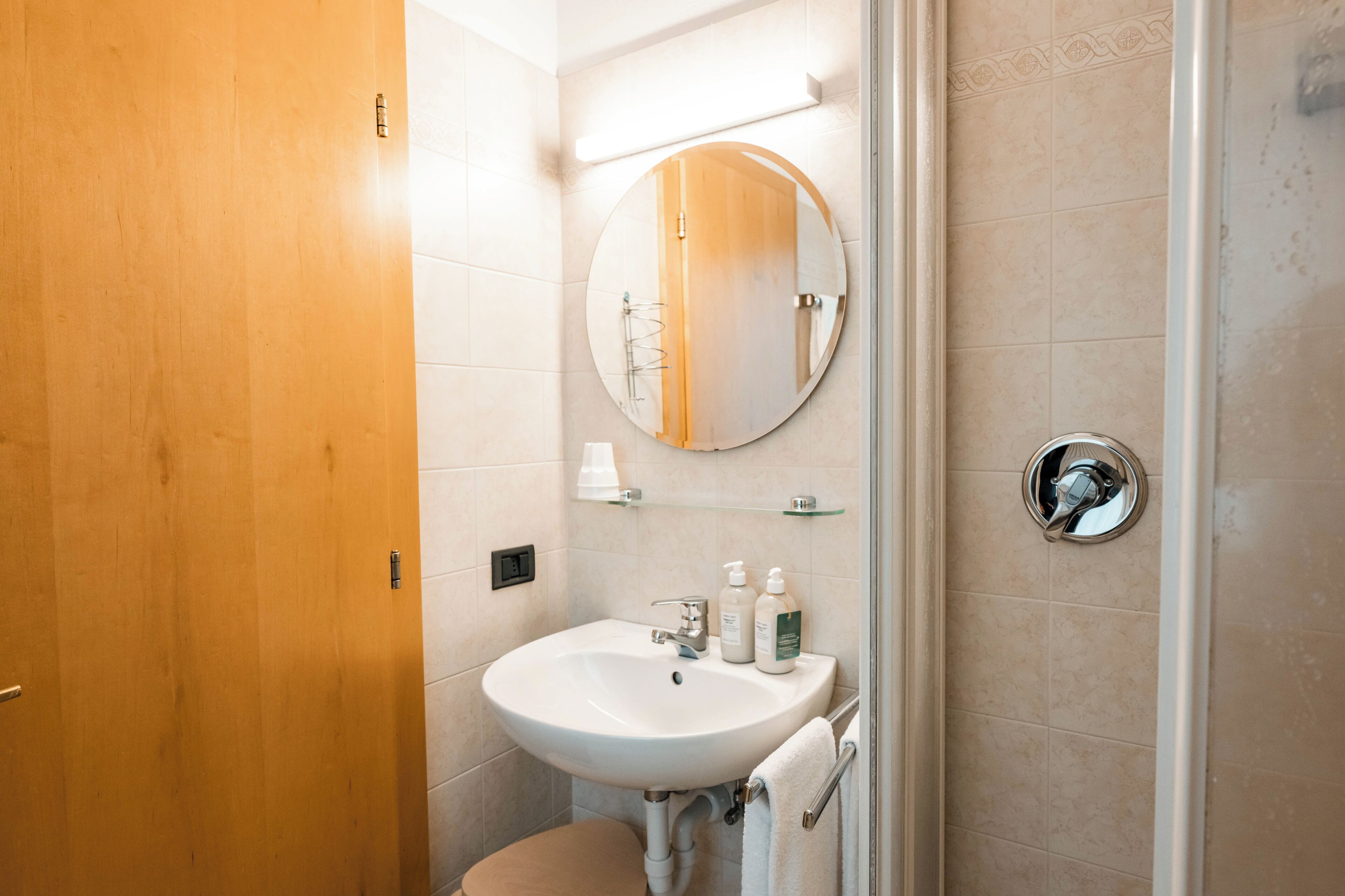 Un baño con puerta de madera, un lavabo blanco, un espejo redondo y zona de ducha.