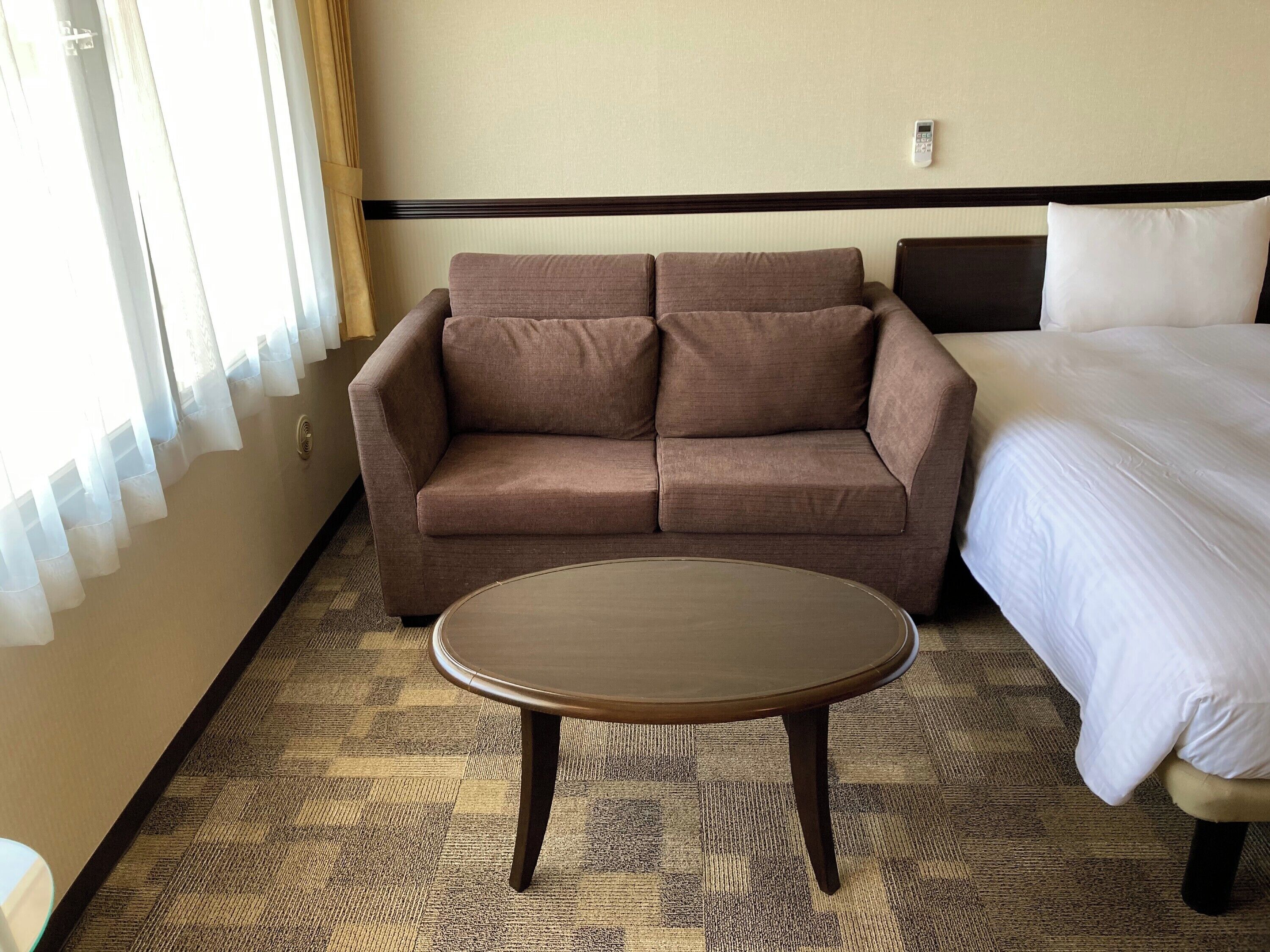 Ein Hotelzimmer mit braunem Sofa, einem runden Tisch, einem Bett mit weißer Bettwäsche und einem Fenster mit durchsichtigen Vorhängen.
