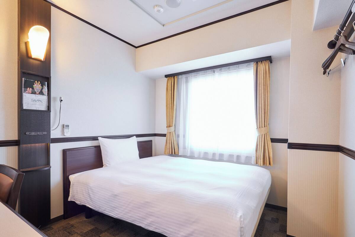 A hotel room with a bed, a desk, a chair, a window with curtains, and a wall-mounted light fixture.