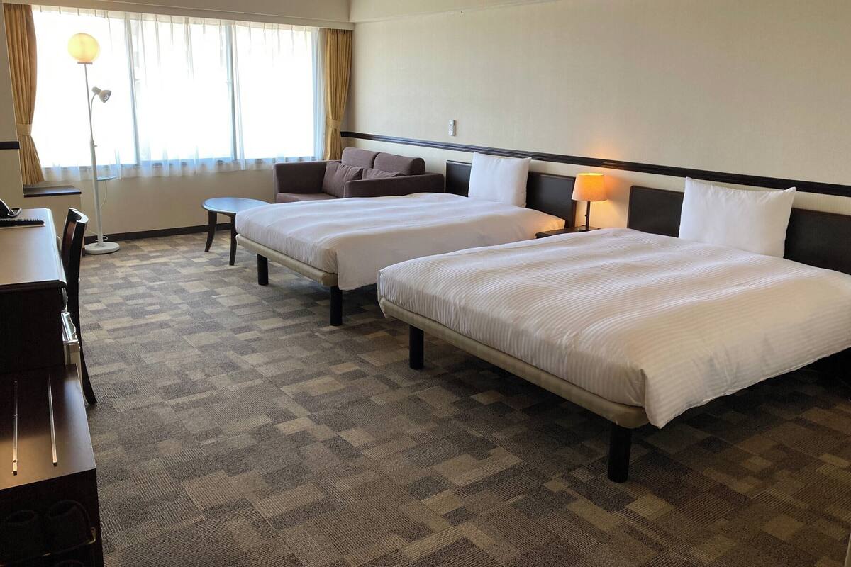 A hotel room with two beds, a desk, a chair, a lamp, and a window with curtains.
