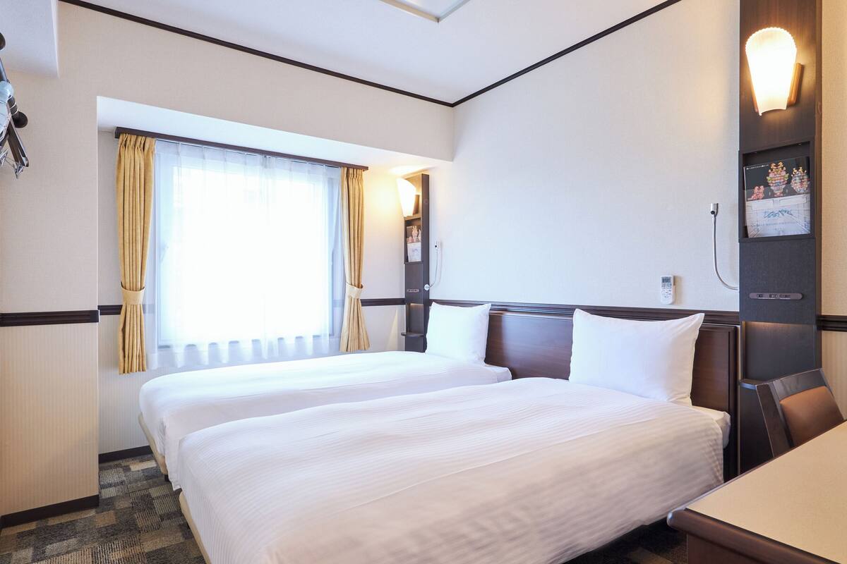 A hotel room with two beds, a large window with curtains, a wall-mounted lamp, and a small desk.