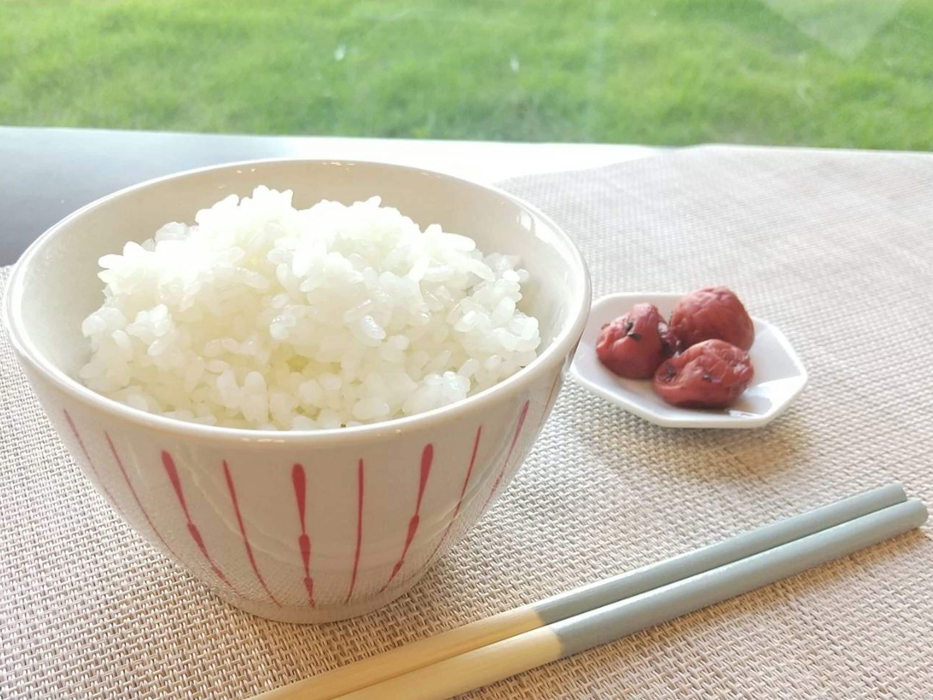 A bowl of white rice with chopsticks and a small plate of red items on a table.