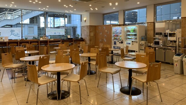 A spacious indoor area with multiple round tables and chairs, vending machines, and a view of a modern building through large windows. - Toyoko Inn Shin Yokohama Station Shinkan (Yokohama)