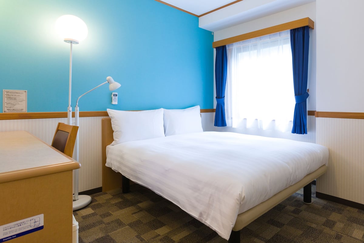 Una habitación de hotel con cama, escritorio, lámpara y ventana con cortinas.