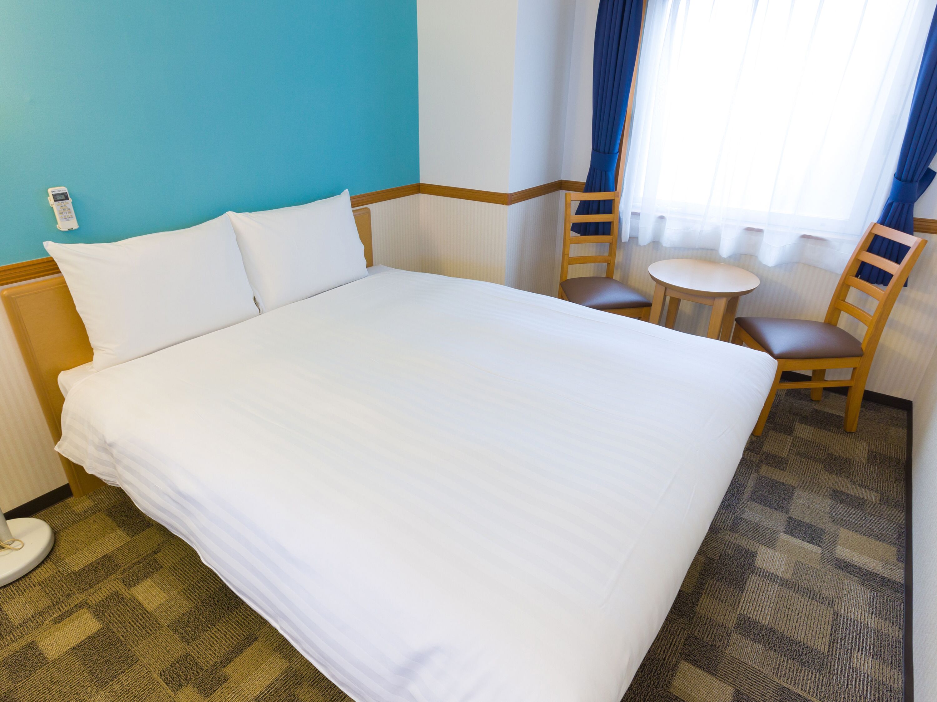 Ein Hotelzimmer mit einem großen Bett, zwei Holzstühlen, einem kleinen runden Tisch und einem Fenster mit Vorhängen.