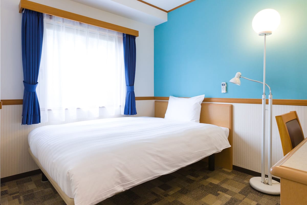 Una habitación de hotel con cama, escritorio, lámpara y ventana con cortinas.