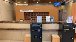 Un espace d’accueil avec un comptoir en bois, deux distributeurs automatiques de billets noirs, une horloge et deux écrans fixés au mur.