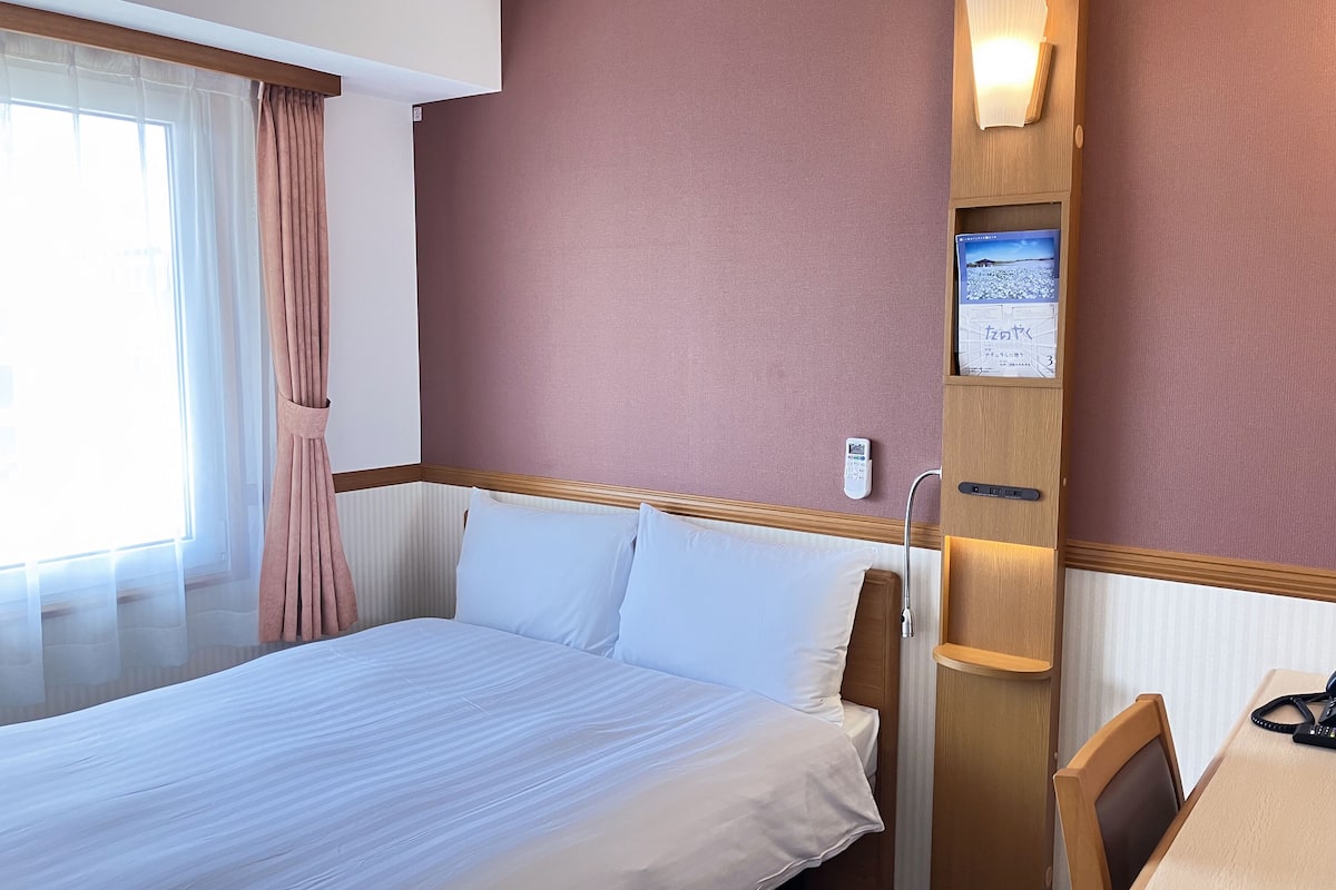Ein Hotelzimmer mit einem Bett, einem Schreibtisch, einem Sessel, einem Nachttisch, einer Lampe, einem Fenster mit Vorhängen und einer wandmontierten Bedieneinheit.