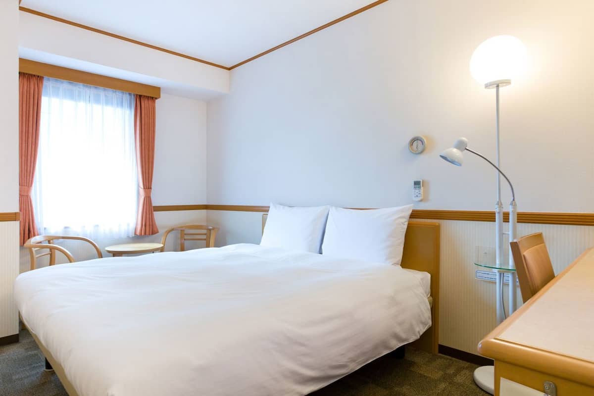 Ein Hotelzimmer mit einem großen Bett, einem Schreibtisch, einem Stuhl, einer Lampe und einem mit Vorhängen versehenen Fenster.