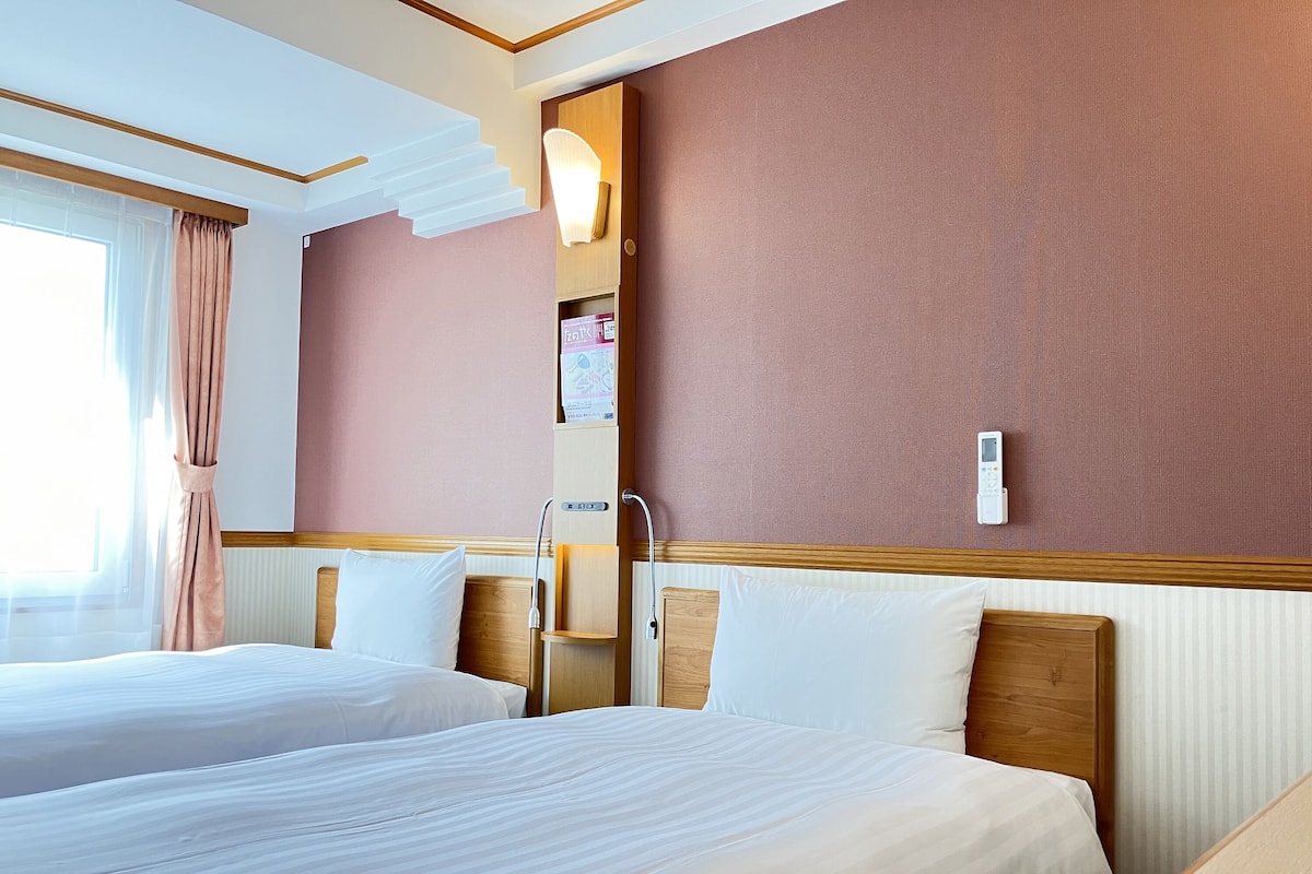 Ein Hotelzimmer mit zwei Betten, einem Holzkopfende, einer wandmontierten Steuerung und einem Fenster mit Vorhängen.
