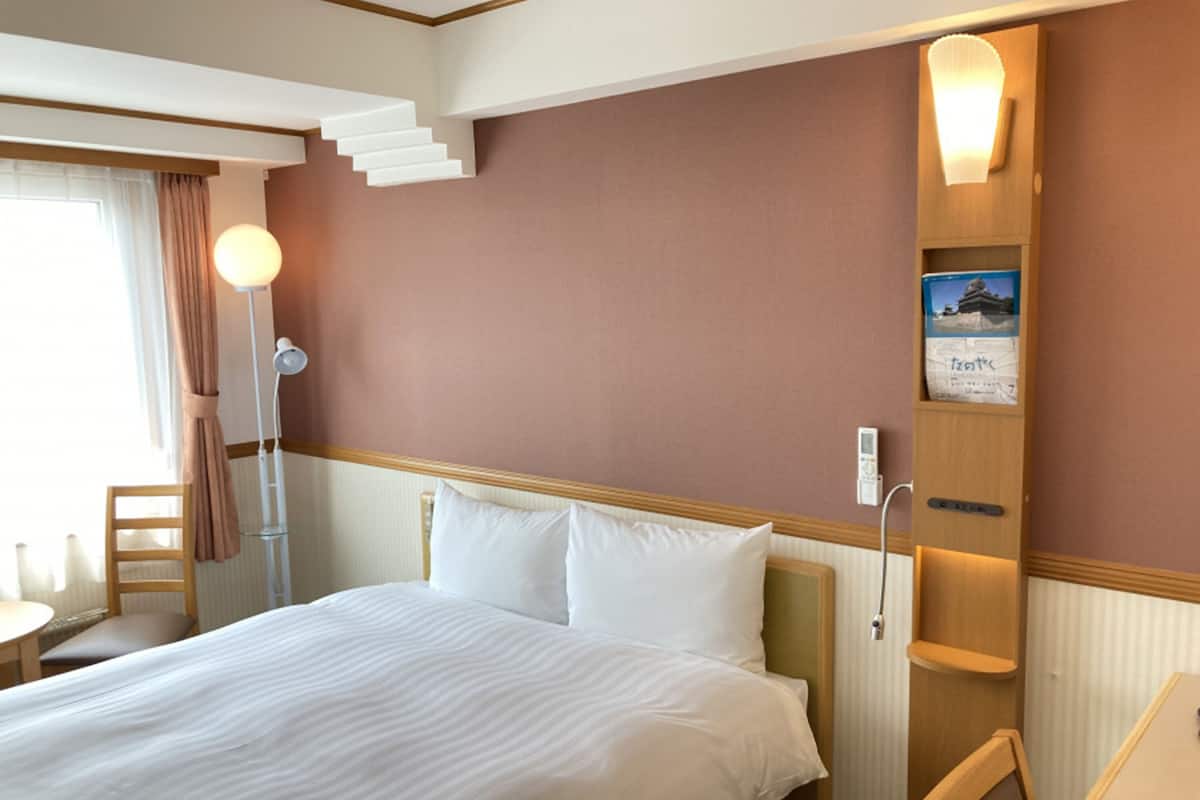 Ein Hotelzimmer mit einem Bett, einem Schreibtisch, einem Sessel, einer Lampe und einem an der Wand befestigten Regal mit einem Buch.