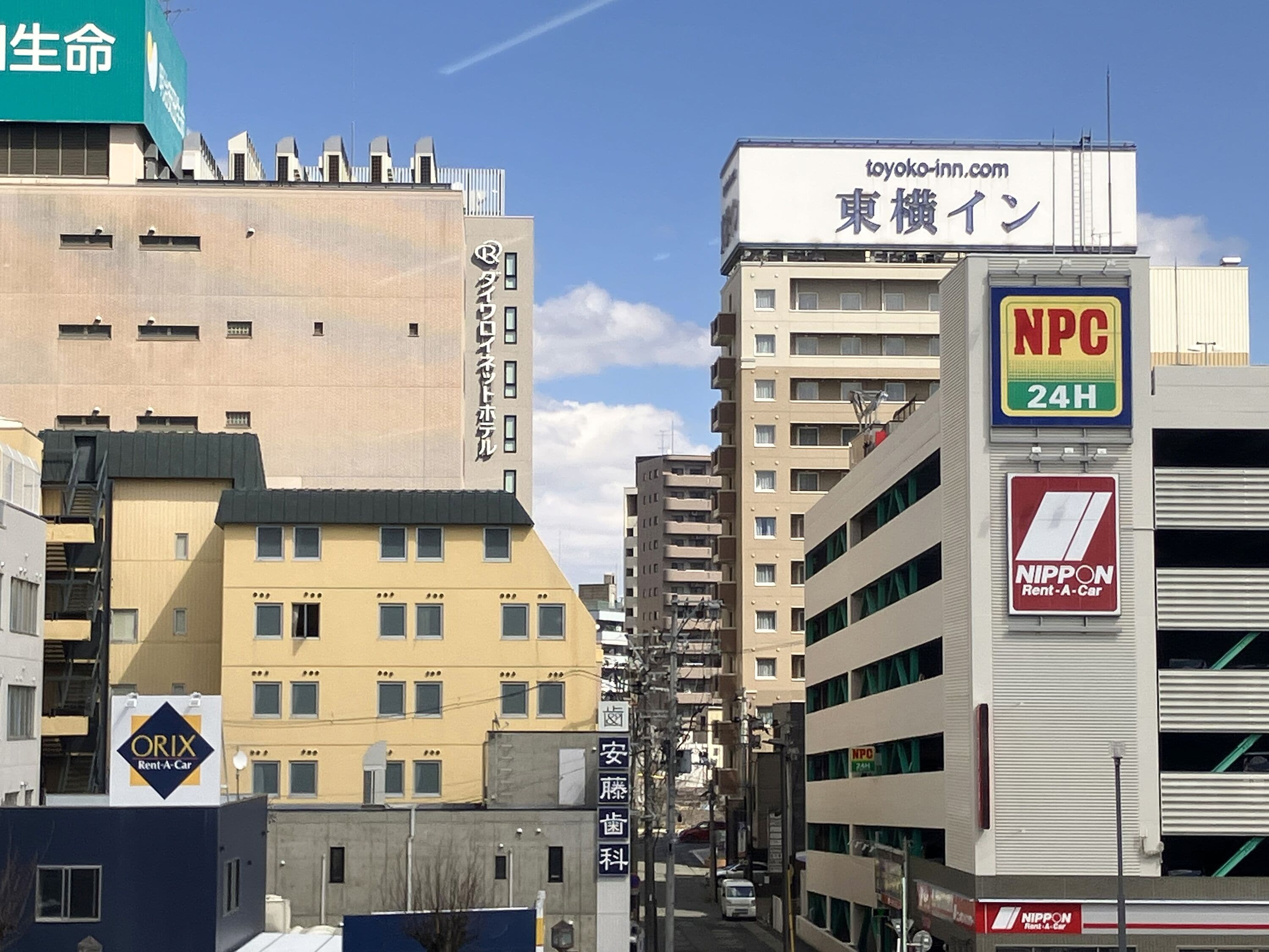 A city street with multi-story buildings, including a sign for a 24-hour convenience store and a rental car service.