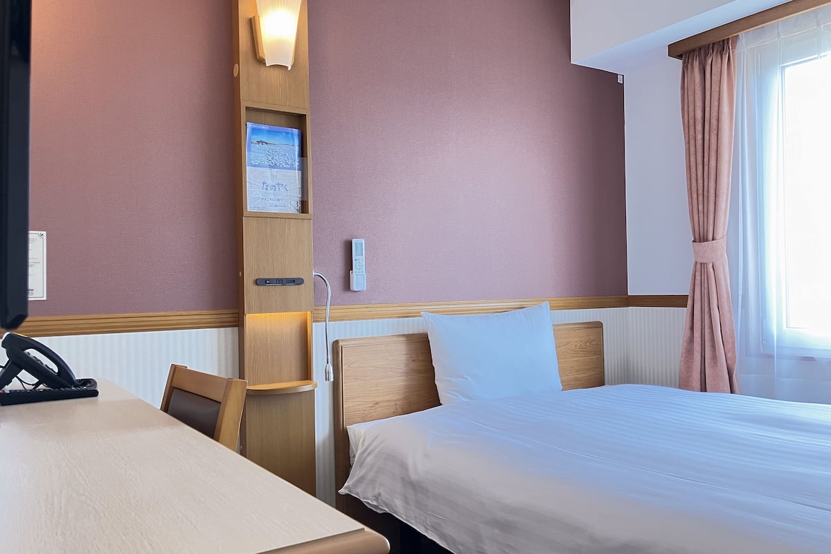 Ein Hotelzimmer mit einem Bett, einem Schreibtisch, einem Stuhl, einer Wandleuchte und einem Fenster mit Vorhängen.