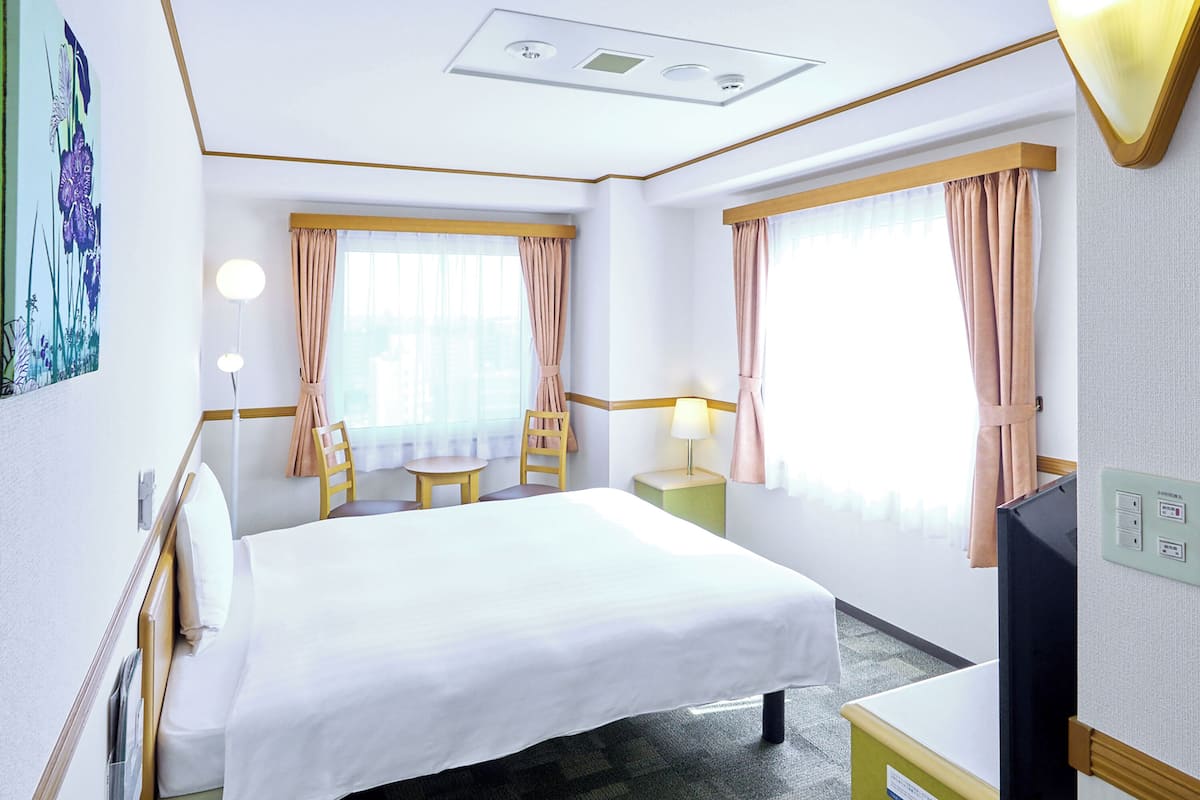 Ein Hotelzimmer mit Bett, kleinem Tisch mit Stühlen, Schreibtisch mit Lampe, Fernseher und einem Fenster mit Vorhängen.