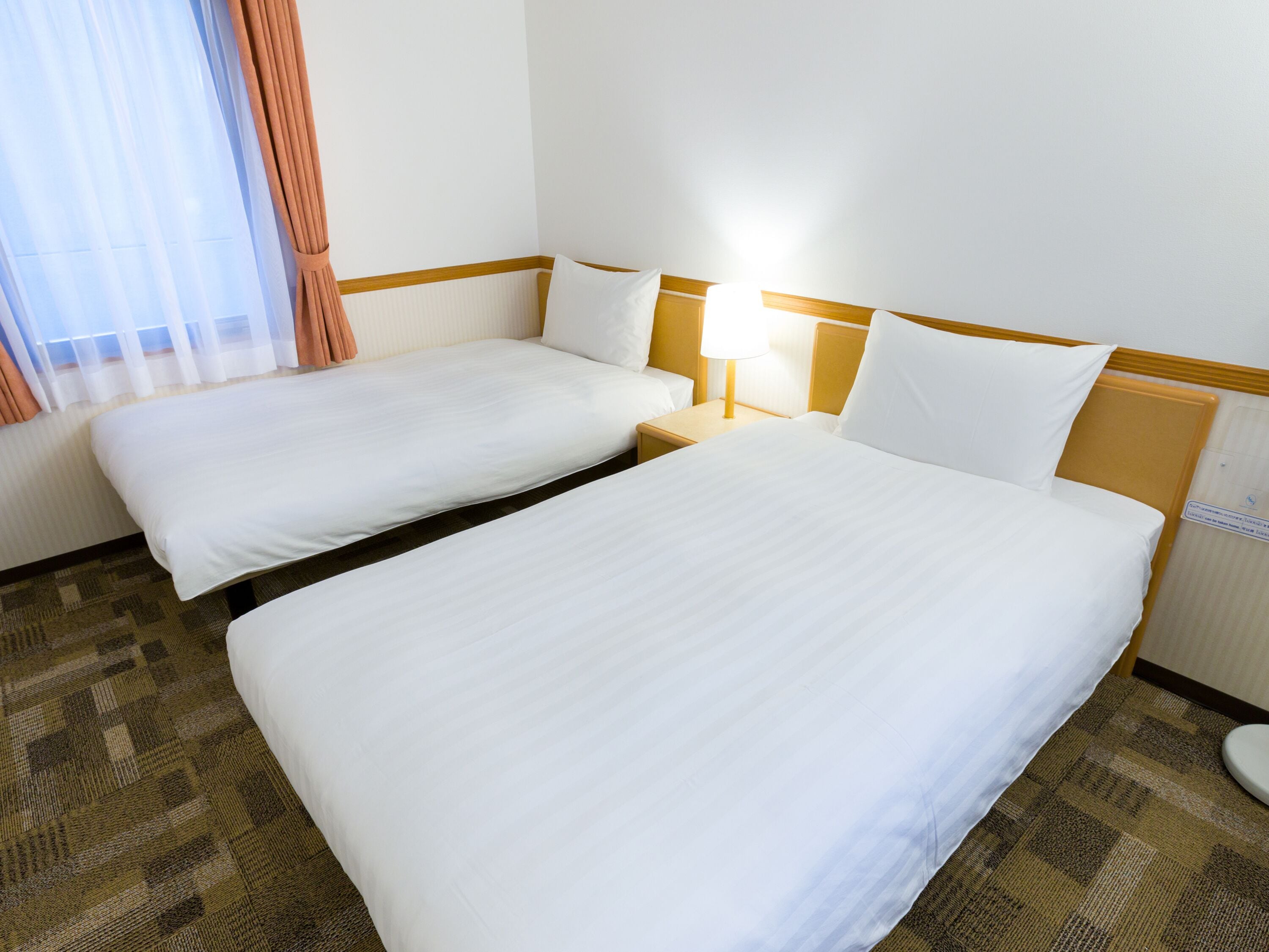 A hotel room with two single beds, a nightstand with a lamp, and a window with curtains.