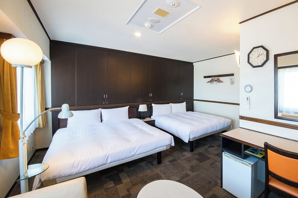 A hotel room with two beds, a desk, a chair, a small refrigerator, and a clock on the wall.