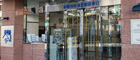 A hotel entrance with a glass door, a sign indicating 'Fukuoka Inn Front Entrance', and a blue carpet leading inside.