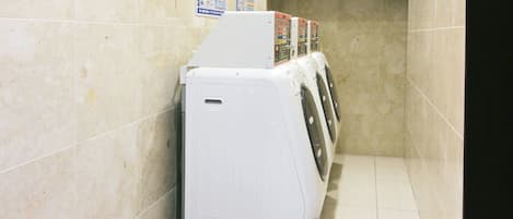 A laundry room with a row of white washing machines and dryers, tiled walls, and a black cabinet above.