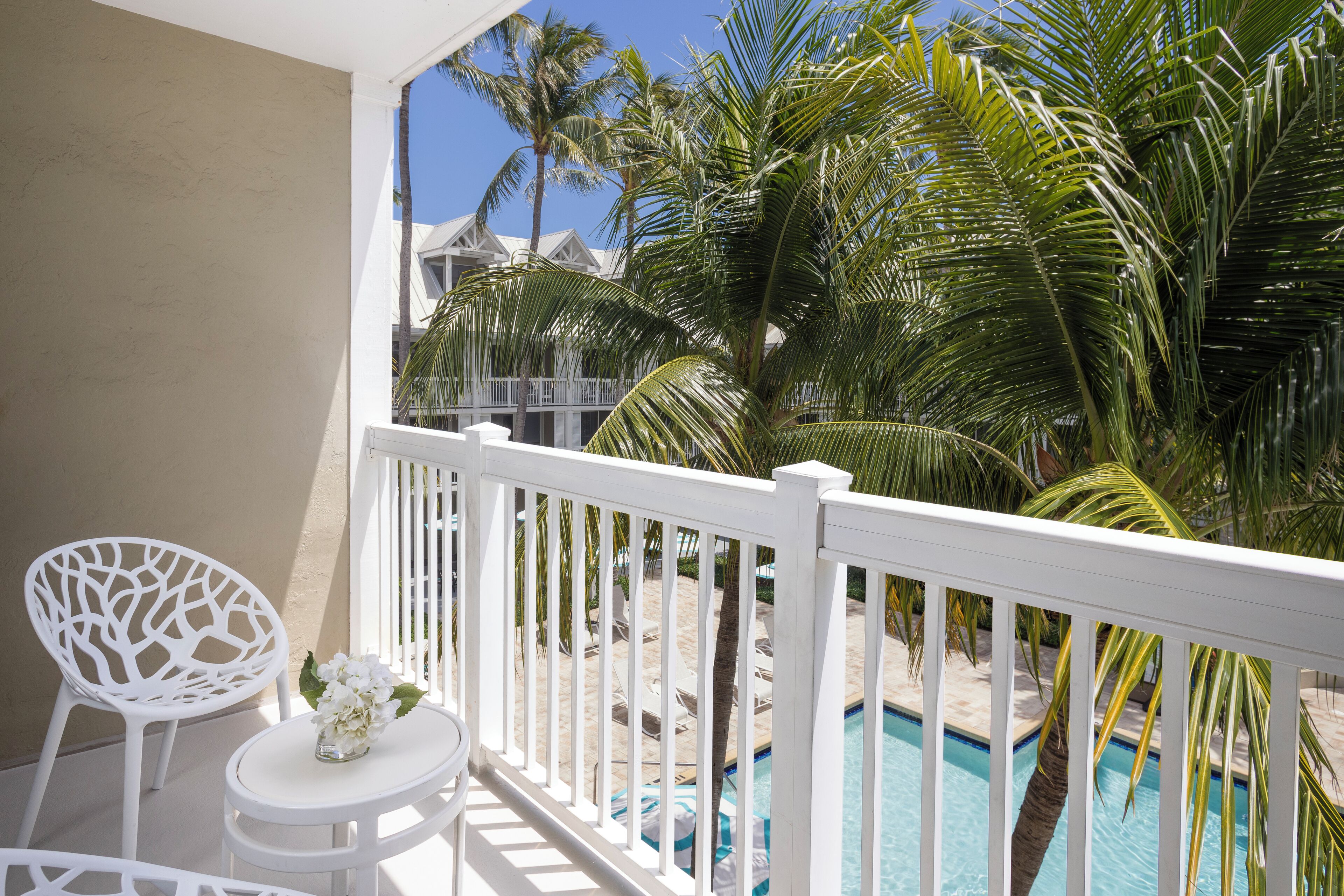 A balcony with white railings, a small table with a vase of flowers, and a pool area with palm trees and lounge chairs.