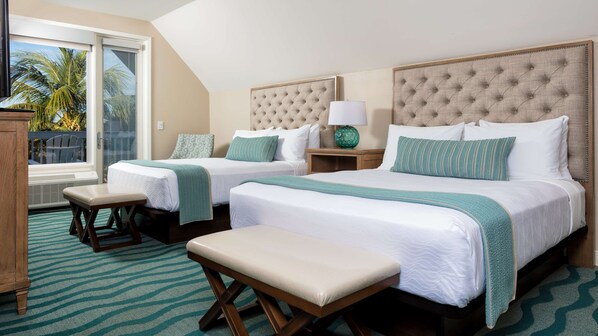Habitación de hotel con dos camas, un banco, una mesita de noche con lámpara y vista a palmeras a través de una puerta corrediza.