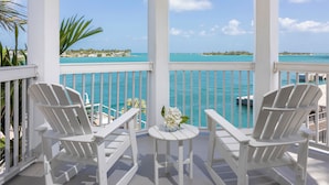 Un balcon abrité, agrémenté de chaises Adirondack blanches et d’une petite table ornée de fleurs, d’où l  ’on peut admirer une mer turquoise et une côte lointaine.
