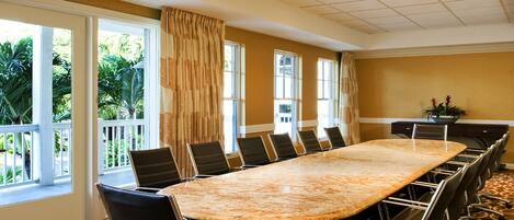 A conference room with a long table and chairs, large windows with curtains, and a view of greenery outside.