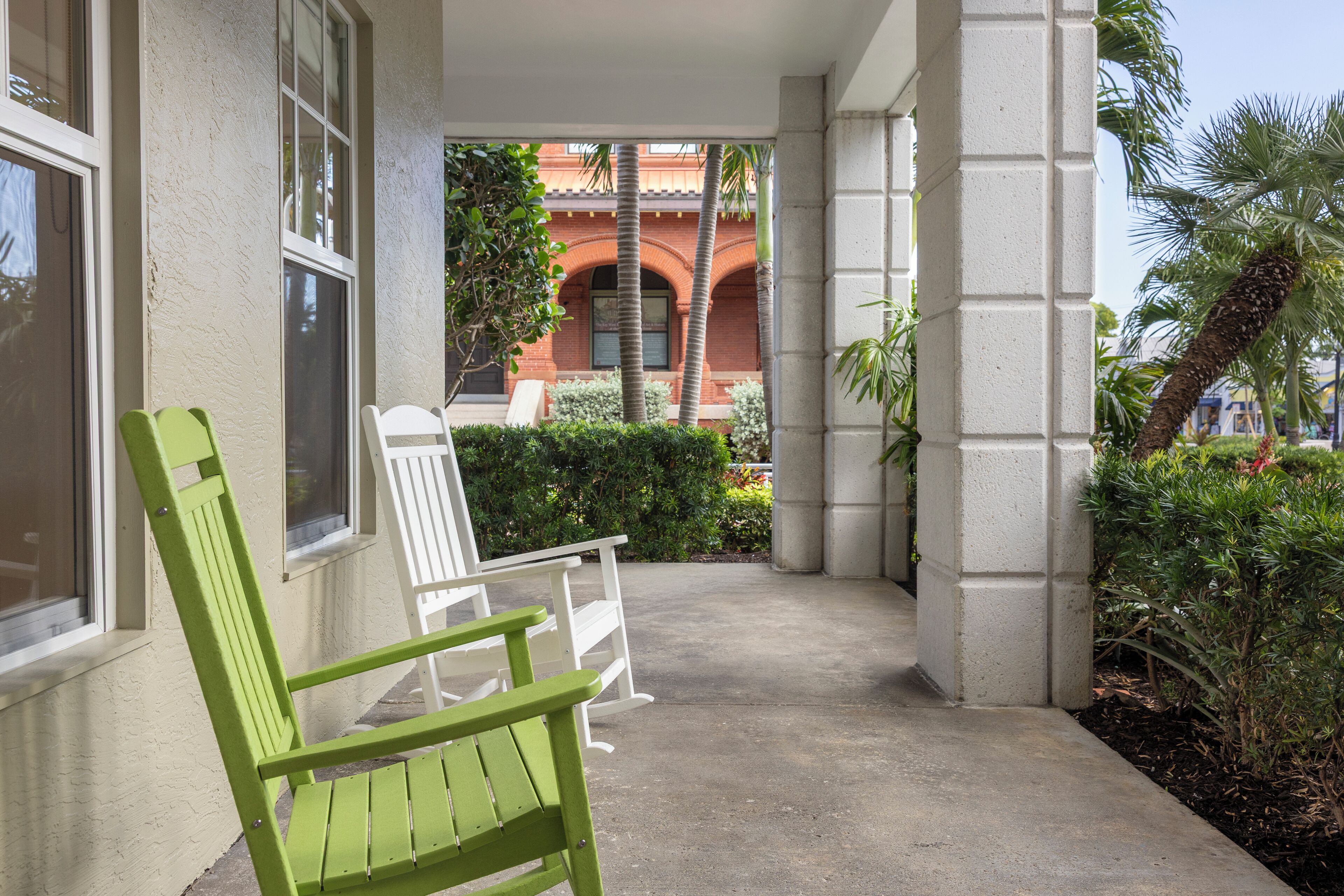 A covered outdoor area with a green chair and a white chair, a concrete floor, and a view of a building with red brick and arches.