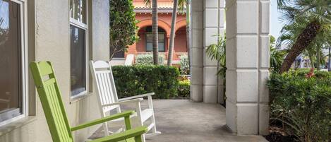 A covered outdoor area with a green chair and a white chair, a concrete floor, and a view of a building with red brick and arches.