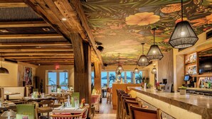 L’intérieur d’un restaurant avec des poutres en bois, un coin bar et une fresque colorée au plafond.