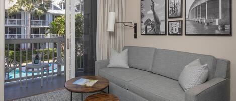Un salon avec un canapé gris, deux petites tables rondes en bois, une grande fenêtre donnant sur une piscine et des immeubles, et des tableaux encadrés accrochés au mur.