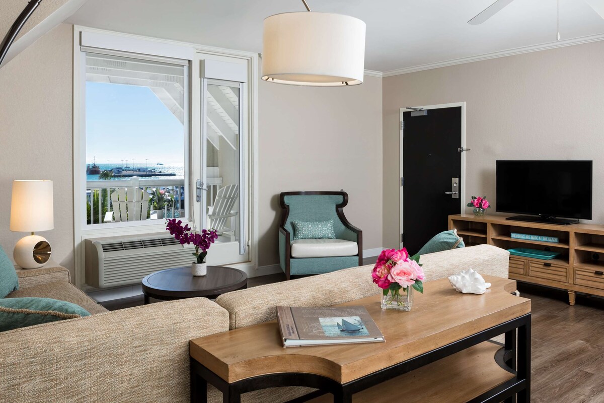 Un salon moderne avec un canapé, une table basse, un téléviseur et une vue sur la mer, visible à travers une grande fenêtre.
